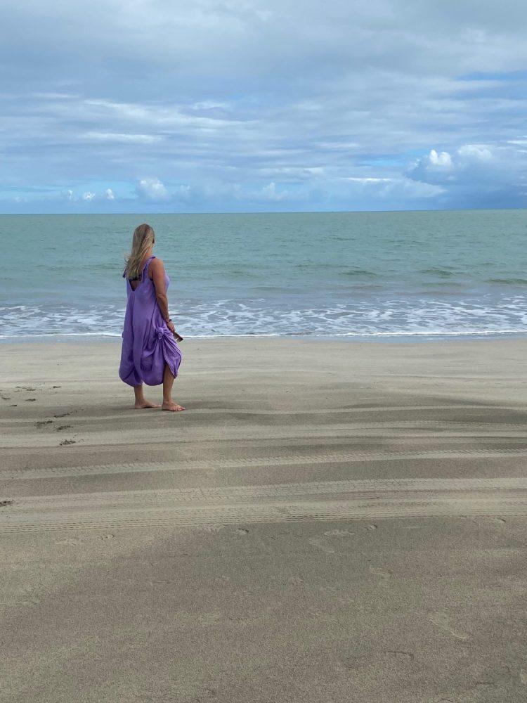 barefoot woman with blonde hair in purple dress standing at a sandy beach looking peaceful out at turquoise ocean vertical view