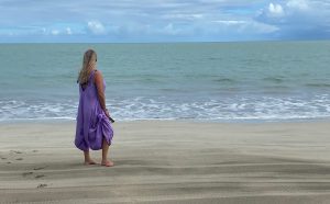 barefoot woman with blonde hair in purple dress standing at a sandy beach looking peaceful out at turquoise ocean horizontal view